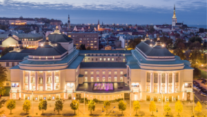 Pildil on vaade Rahvusooper Estonia hoonele õhtusel ajal, kui teatri fassaadil on sisse lülitatud välisvalgustus. Hoone on helekollast värvi ja roheka katusega.