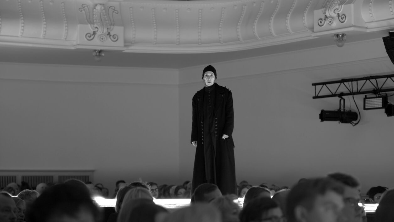 Mustvalgel fotol on stseen lavastusest "Macbeth". Pildi alaosas on näha publikuliikmete päid, keskel seisab pika tumeda rüü ja tumeda mütsiga Kaie Mihkelson aadlidaami rollis.