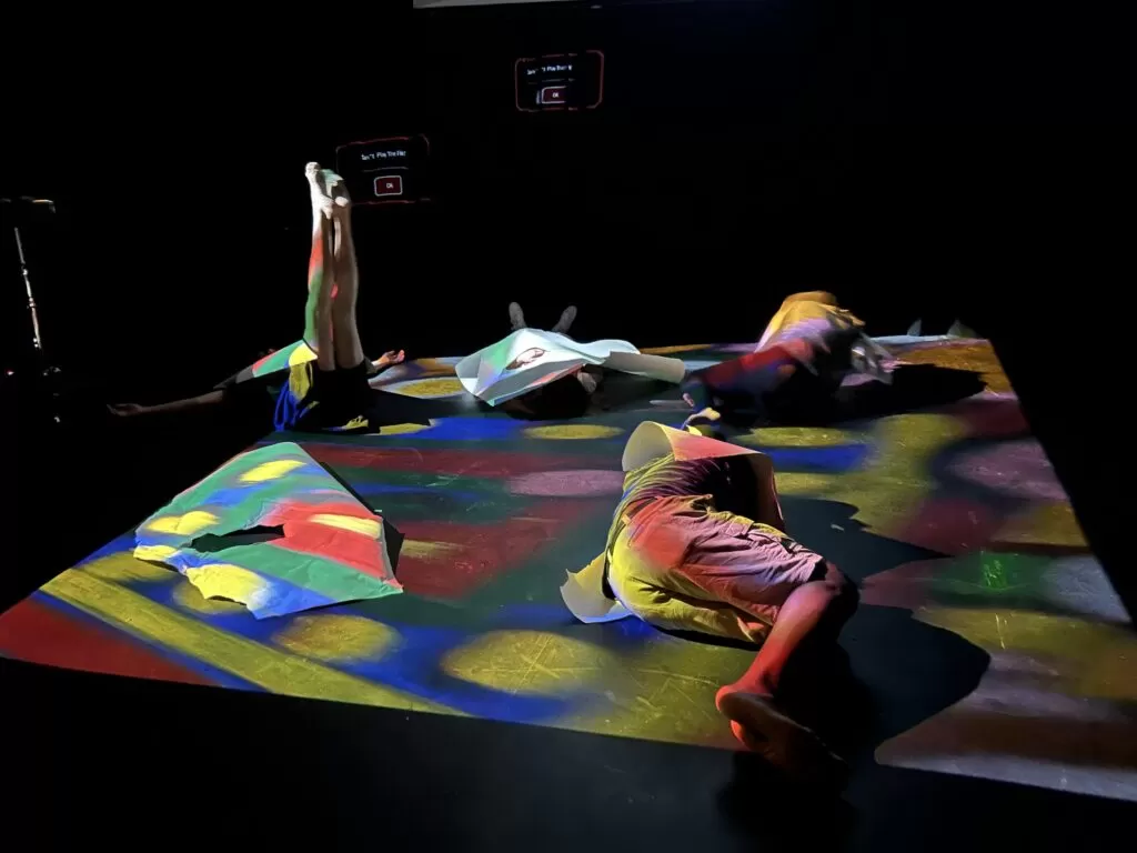 Pildil on neli inimest, kes lamavad musta värvi põrandal, ning nende peale on projitseeritud värvililine ringidega kujutis.