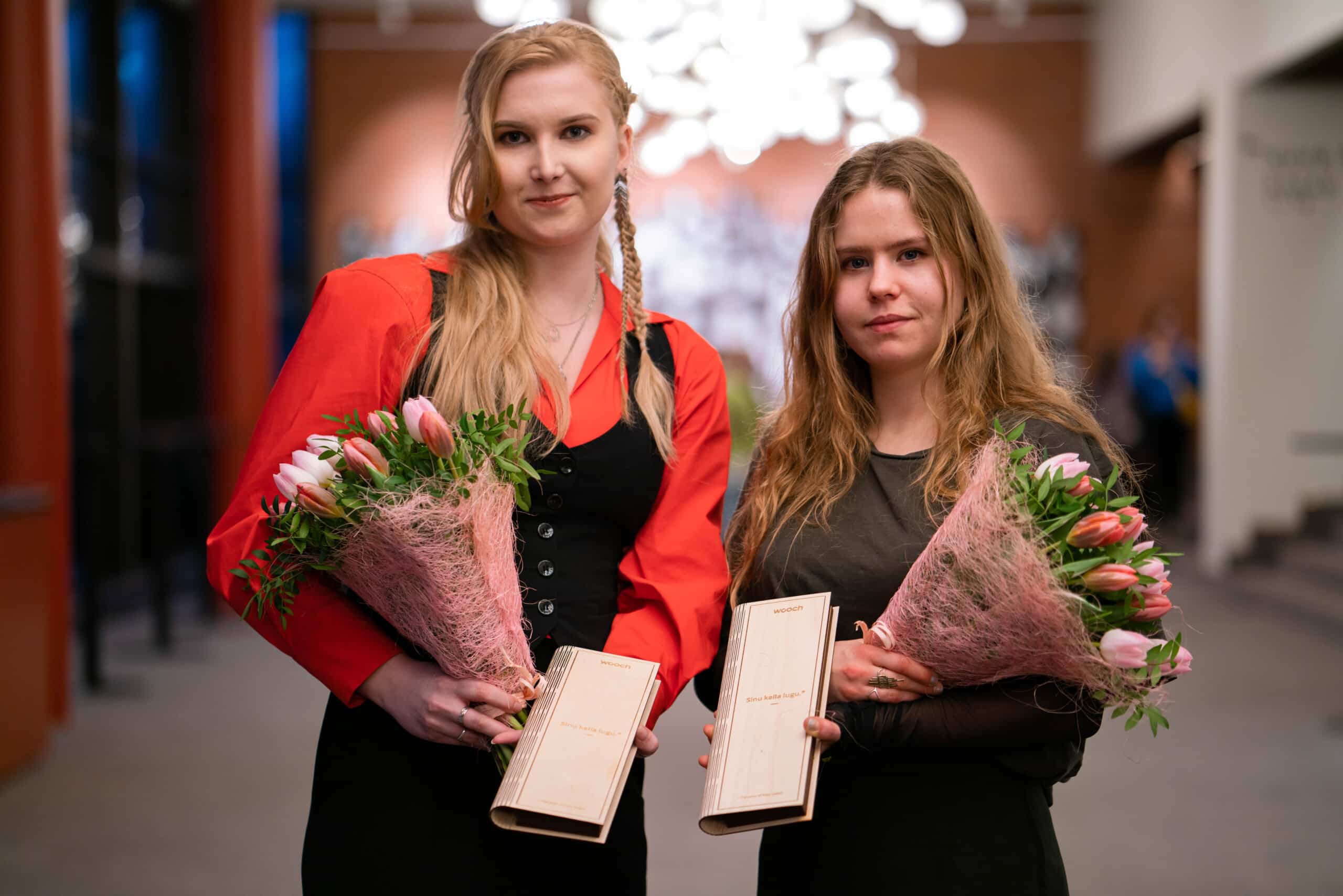 Lysette Tint ja Lisette-Marie Viilup seisavad kõrvuti Ugala Teatri fuajees, mõlemal käes lillekimp ja heledast puidust karp Woochi käekellaga.