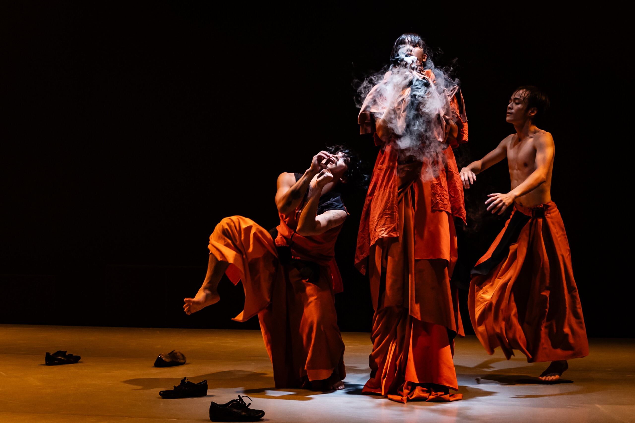 Pildil on kolm tantsijat, riietatud oranžidesse rohke kangaga seelikutesse ja kleitidesse. Kaks liiguvad paljajalu mööda lava, üks seisab näoga valguse poole ning puhub valget suitsu.