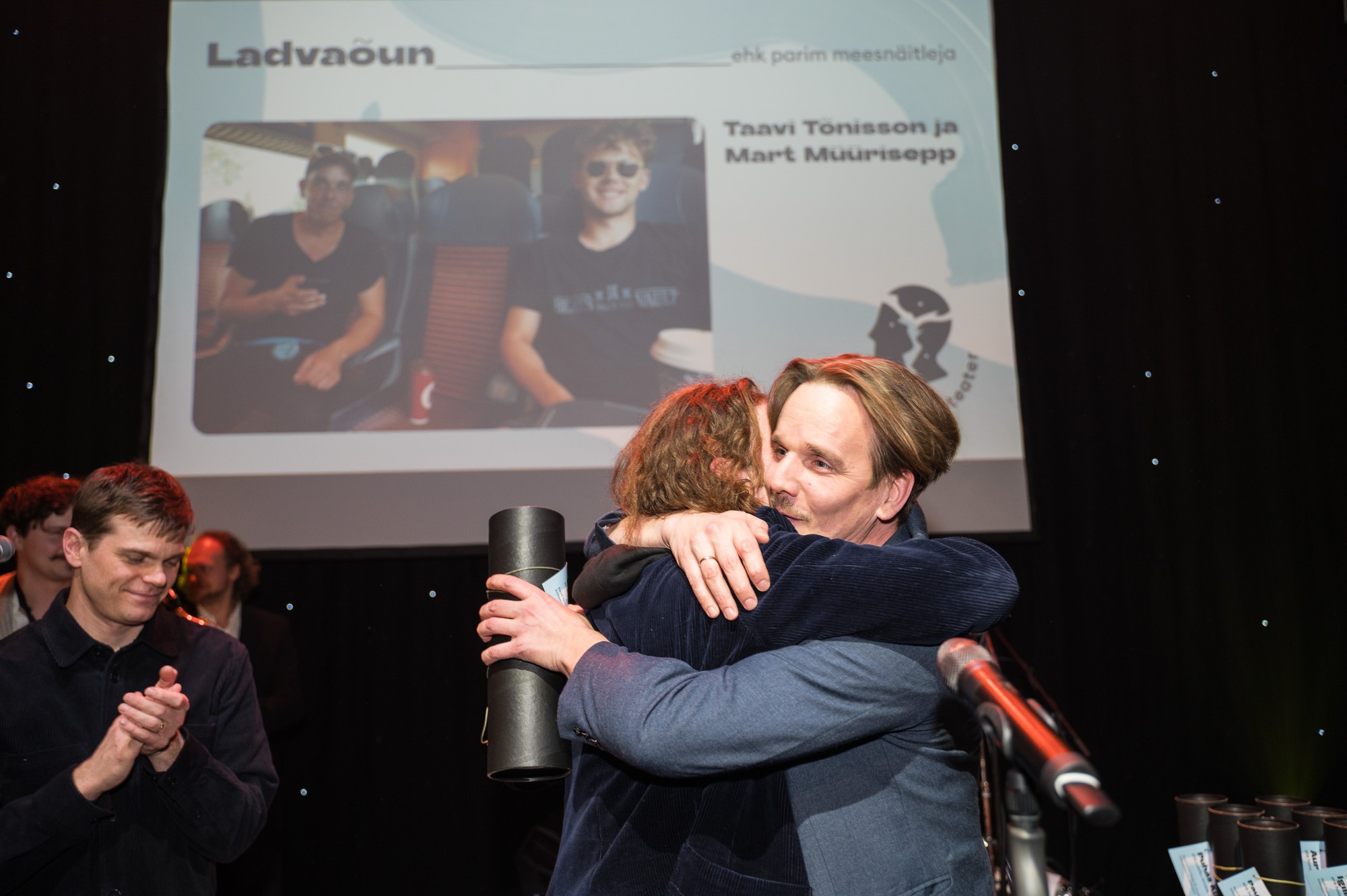 Pildi esiplaanil on Mart Müürisepp ja Taavi Tõnisson, kes kallistavad üksteist. Vasakul eemal seisab Mirko Rajas. Taustal paistab ekraan pildiga Müürisepast ja Tõnissonist.