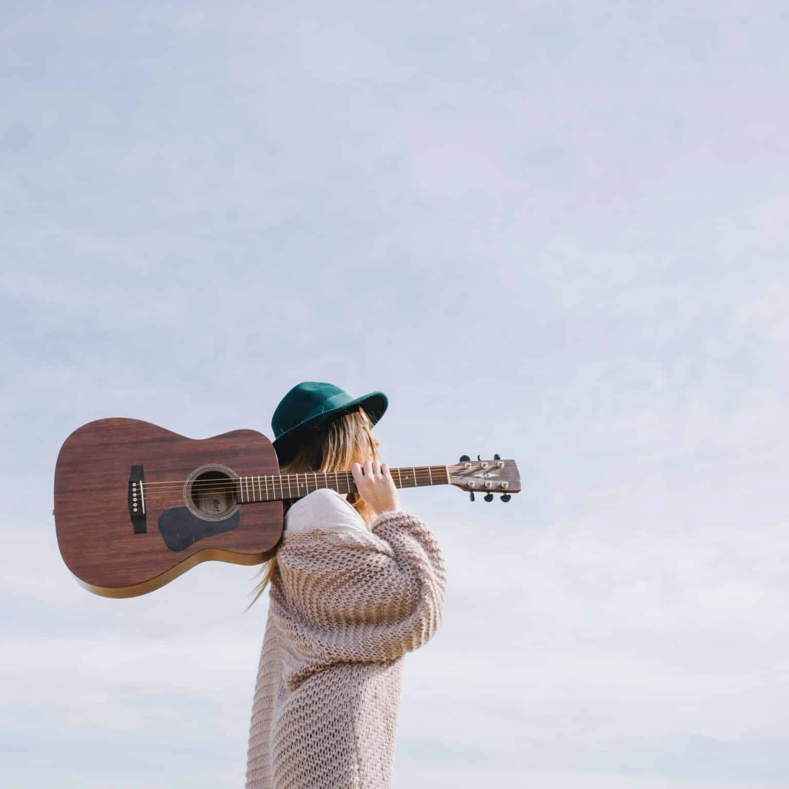 Pildil on naine, kes hoiab õla peal tumepruuni akustilist kitarri. Tal on seljas beež kampsun ja peas rohekas kübar. Taustal paistab helesinine taevas.