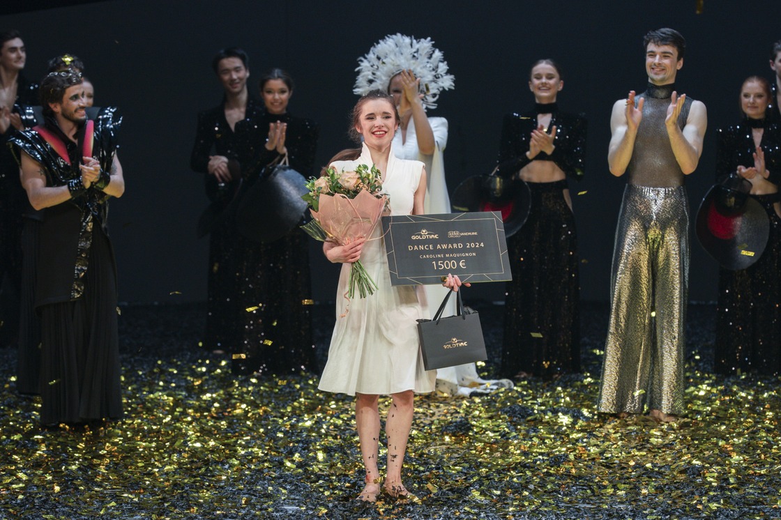 Pildil on Caroline Maquignon, kes seisab laval, seljas valge põlvedeni ulatuv kleit ja käes lillekimp, must Goldtime'i kinkekott ja auhinnadiplom. Teda ümbritsevad kuldsed paberitükid ja taustal seisavad plaksutavad tantsijad.