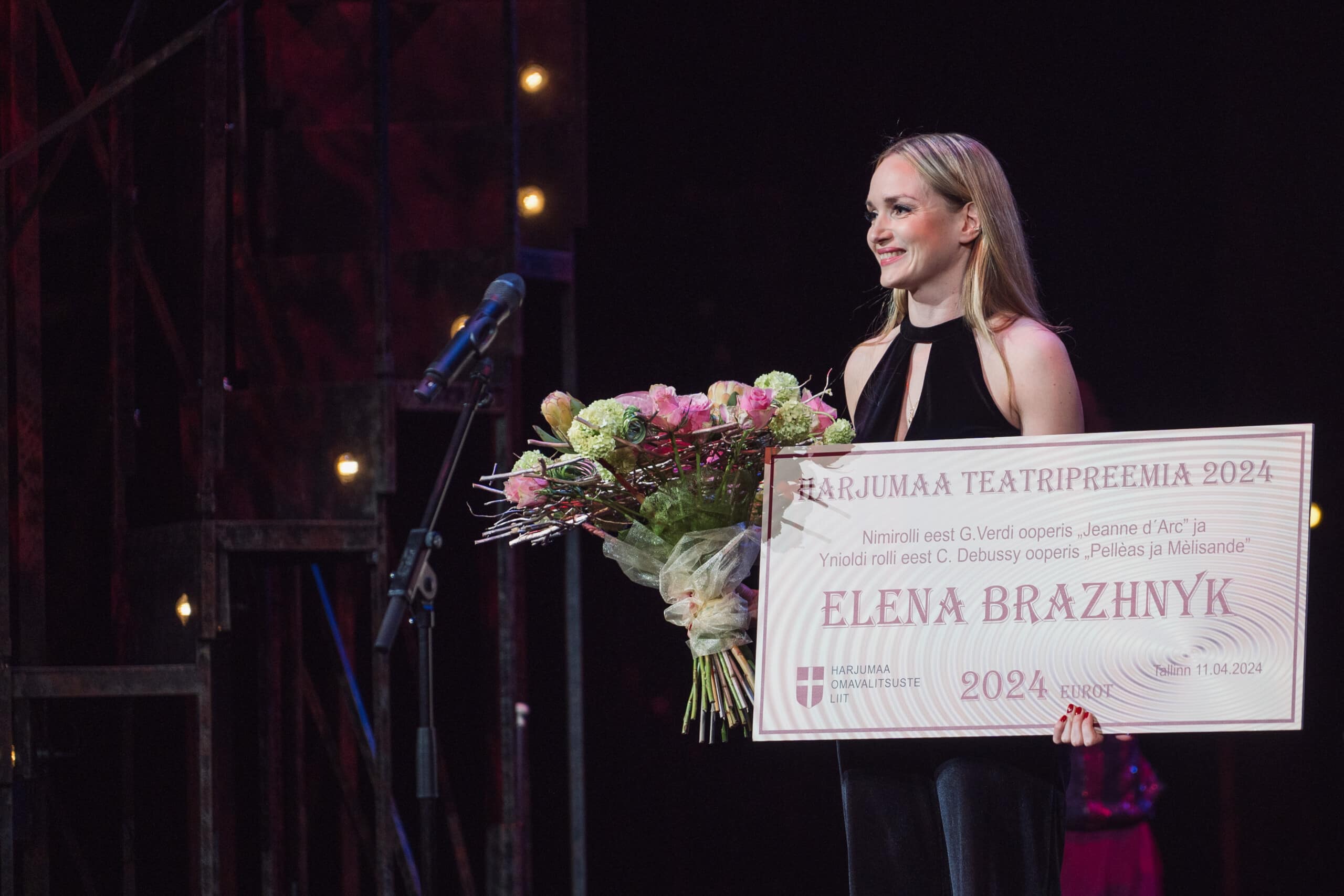 Pildil on Elena Brazhnyk, kes seisab laval, käes lillekimp ja suur diplom. Ta on noor heledate pikkade juustega naine, seljas must varrukateta pluus ja jalas mustad püksid.