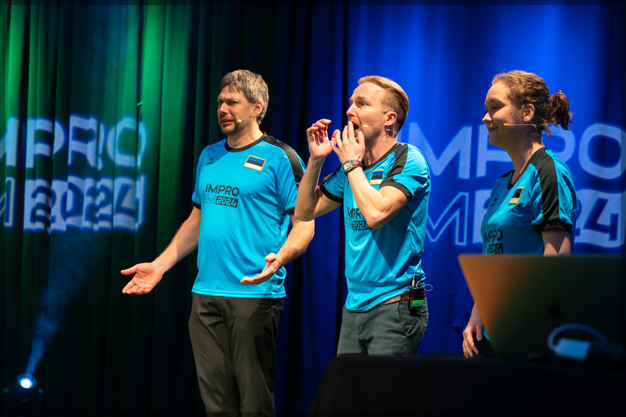 Pildil on Eesti impronäitlejad Maarius Pärn, Rauno Kaibiainen ja Rahel Otsa, kes seisavad laval, seljas sinised väikese Eesti lipuga spordisärgid. Kardinale nende selja taga on projitseeritud "IMPRO EM 2024".