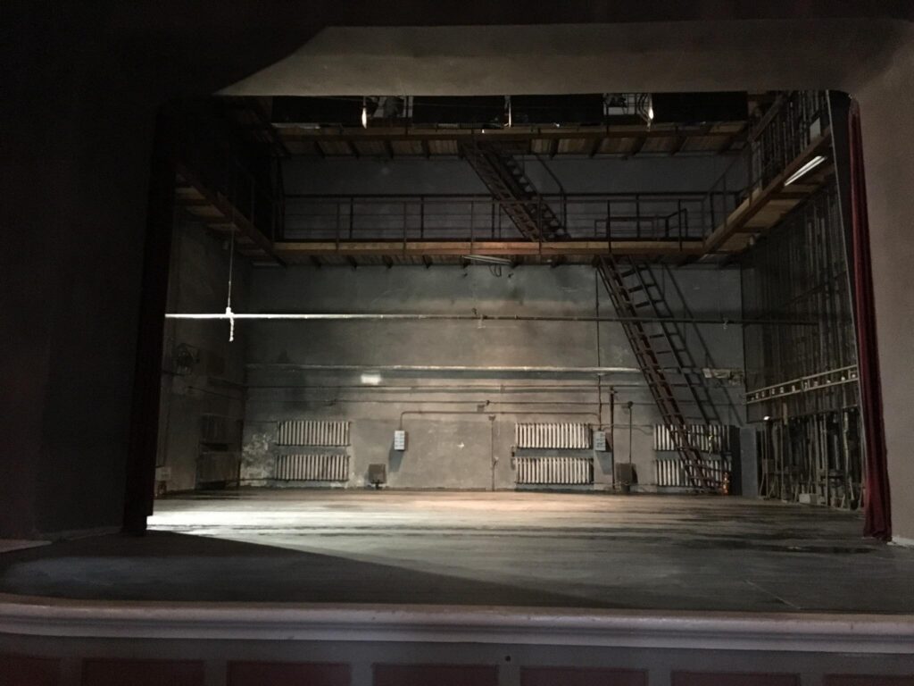 Pildil on Koidu seltsimaja lava, mille seinad ja põrand on tumedad. Tagumises seinas paistavad kuus suurt radiaatorit ning kaks metallist treppi.