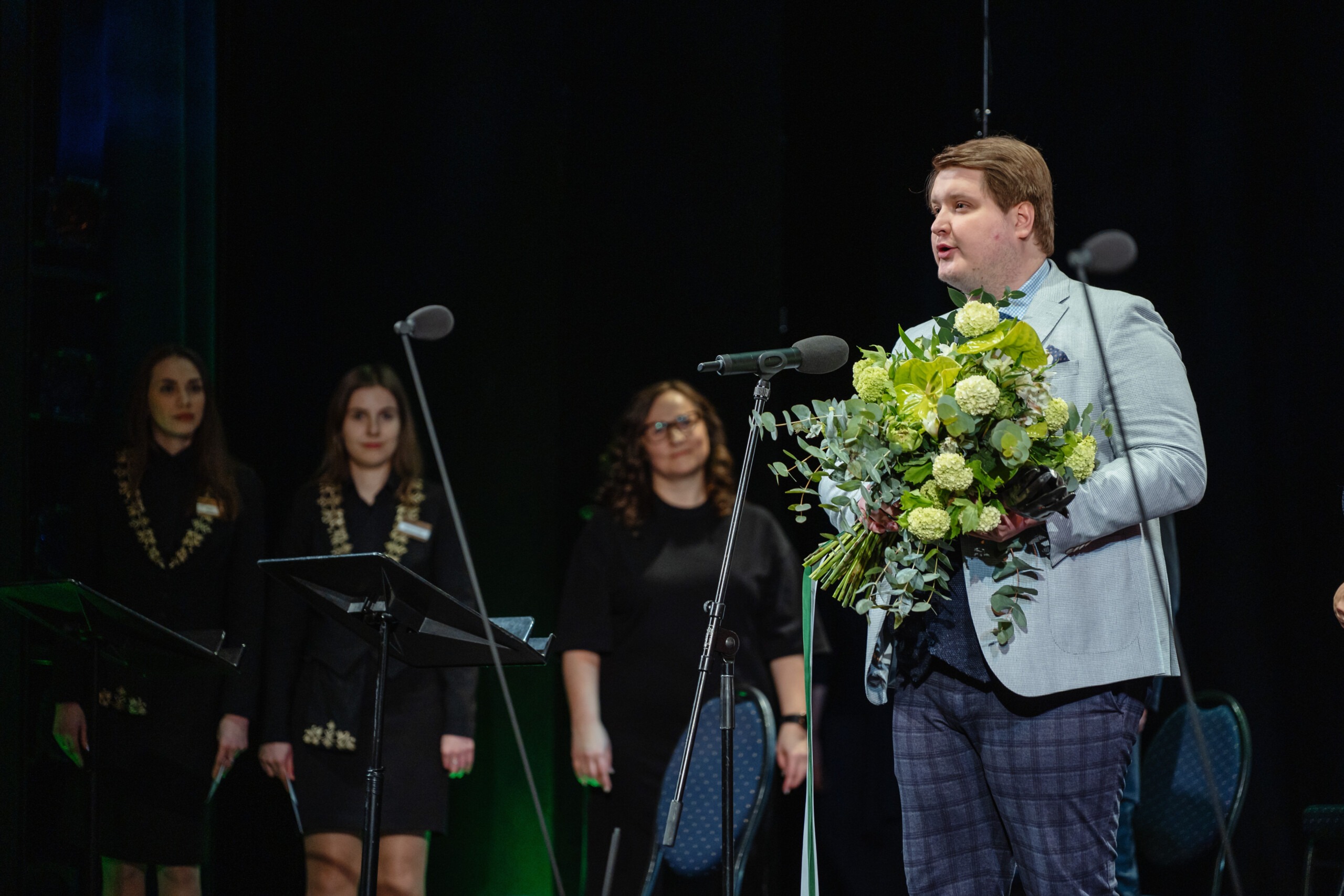 Pildil on Reigo Tamm publikupreemia üleandmisel. Ta seisab laval mikrofoni taga, käes rohekates toonides lillekimp, seljas hall pintsak ja ruudulised püksid. Tema taga paistab kolm noort naist.