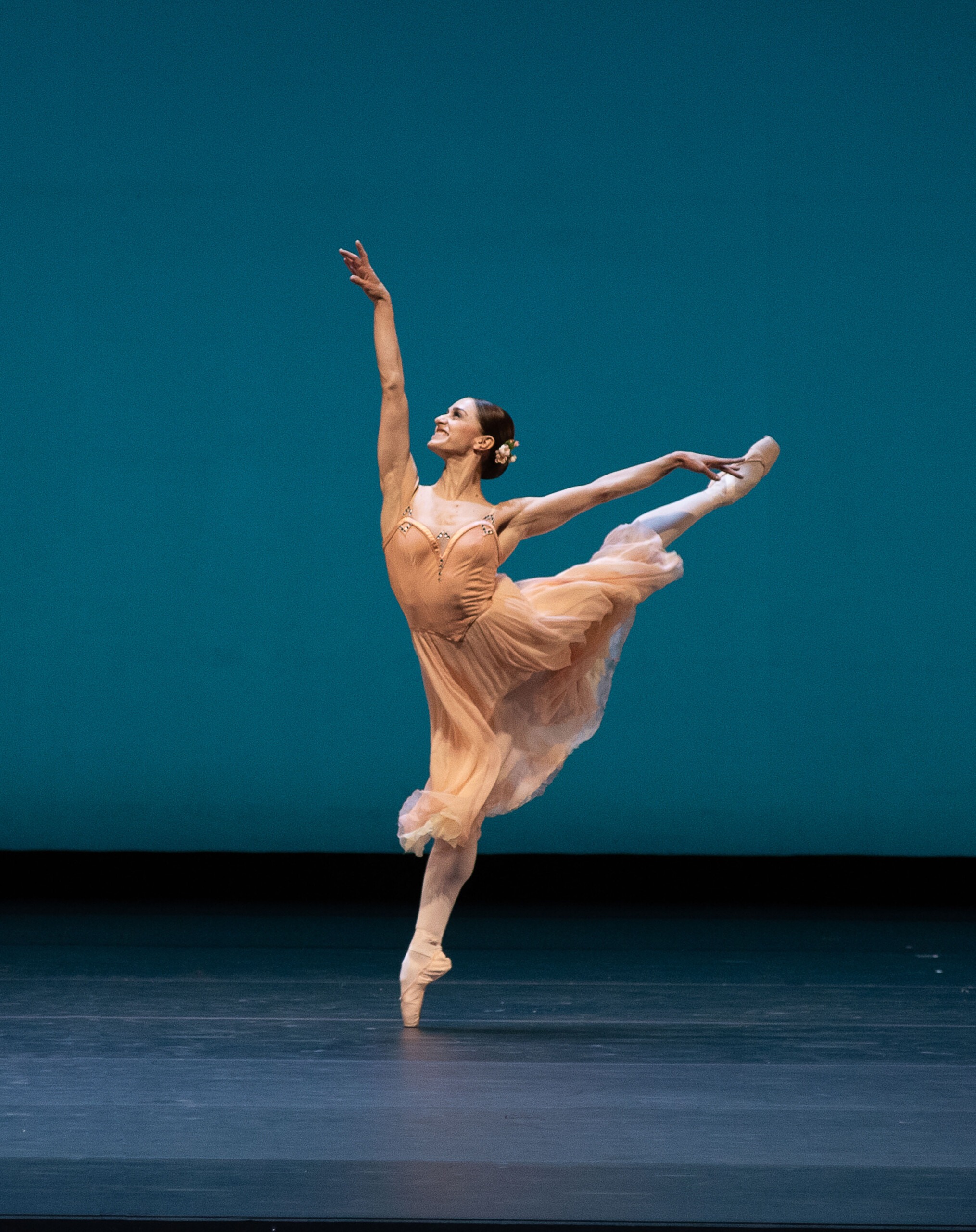 Pildil on baleriin Marianela Núñez, kes seisab laval parema jala varvastel, vasak jalg ja käed ülespoole sirutatud. Tal on seljas õhuline helebeež kleit.
