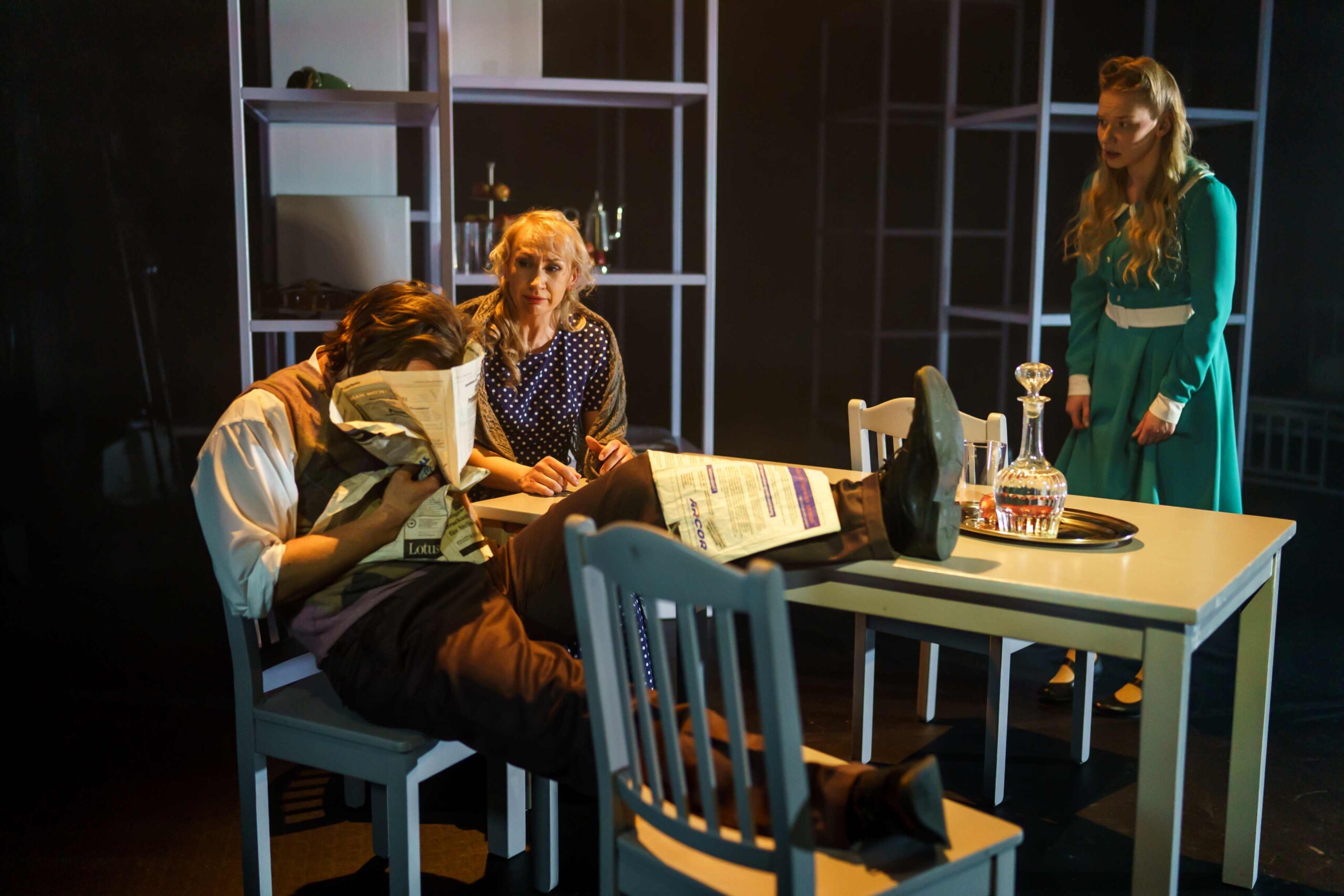 Pildil on stseen Rakvere Teatri lavastusest "Metamorfoos", kus Madis Mäeorg ja Silja Miks istuvad valge laua taga, eemal seisab sinakas kleidis Jaune Kimmel. Mäeorg toetab ühte jalga lauale, teist toolile ning on näo peitnud ajalehe sisse.