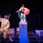 Pildil on lava kahe näitlejaga, üks neist seisab kõrge postamendi peal, käes suur punane pall ja seljas läikiv keep. Teine seisab eemal ja viskab õhku sädelust.