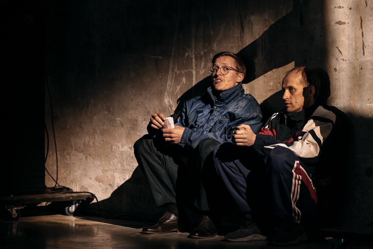 Pildil istuvad hämara betoonseina ääres näitlejad Tõnis Niinemets ja Markus Luik. Neil on seljas 90ndate stiilis kiledressid.