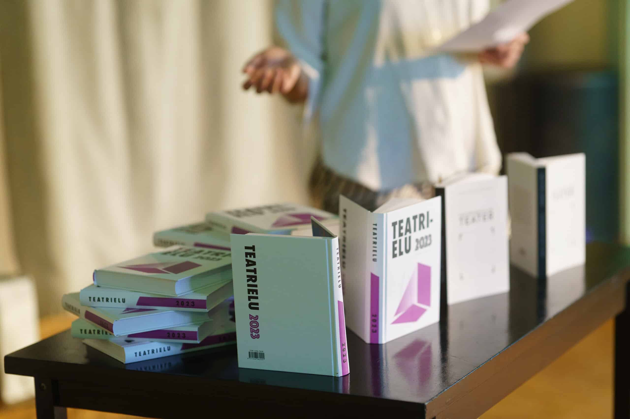 Pildil on musta värvi laud, millel seisab eri asendites rohkelt Teatrielu 2023 raamatuid. Laua taga seisab valges särgis naine.