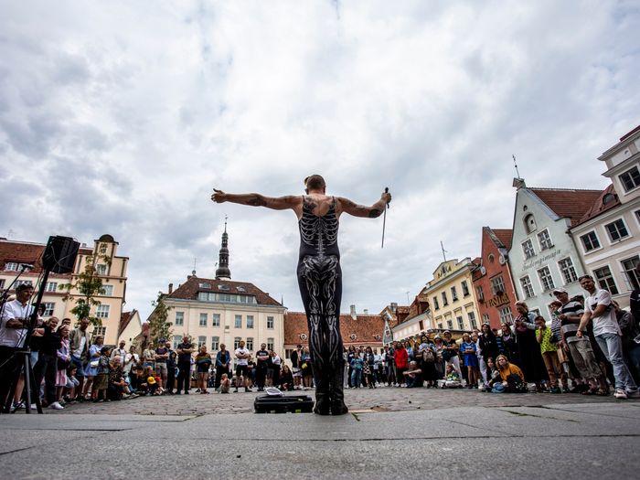 Pildil on etendaja, kes seisab Tallinna Raekoja platsil publiku ees. Tal on seljas must skeletikujutisega kostüüm.