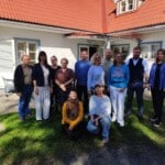Pildil on 12 inimest, kes seisavad ja kükitavad valge puumaja ees. Nende peale langeb puude vari ja nende ümber paistab muru.