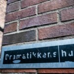 Pildil on telliskividest sein, millele on kinnitatud metallist silt. Sildil on mustal taustal kirjas "Dramatikkens hus".