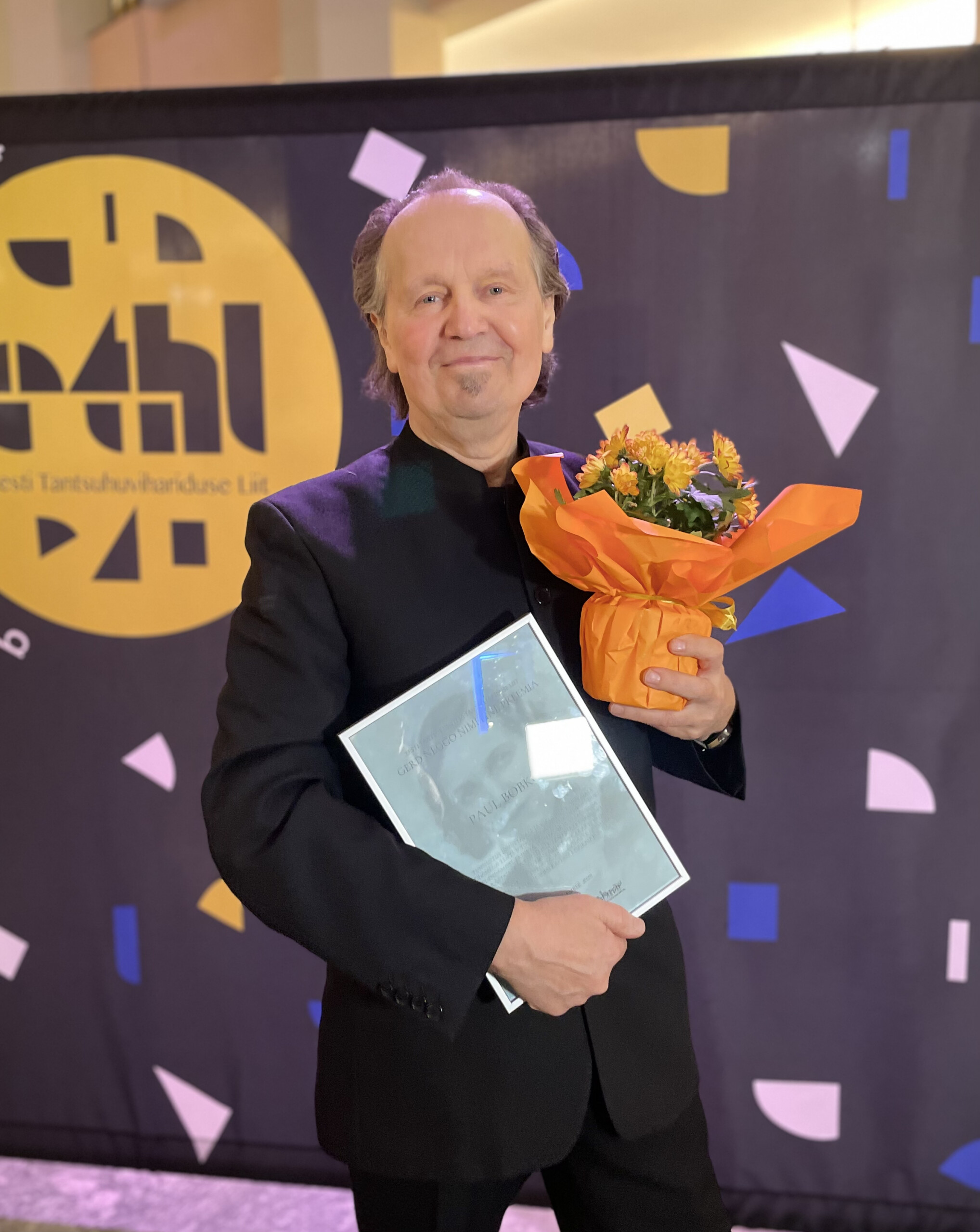 Pildil on Paul Bobkov. Tal on heledad õhukesed ja lühikesed juuksed ning seljas must ülikond. Vasakus käes hoiab ta oranžis ümbrises potilille ja paremas käes raamitud diplomit.