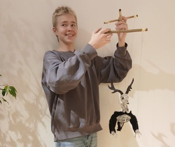 Pildil on heledate juuste ja halli pusaga noormees, kes hoiab käes marionettnukku. Marionett on musta ja valget värvi ning tumedate sarvedega.
