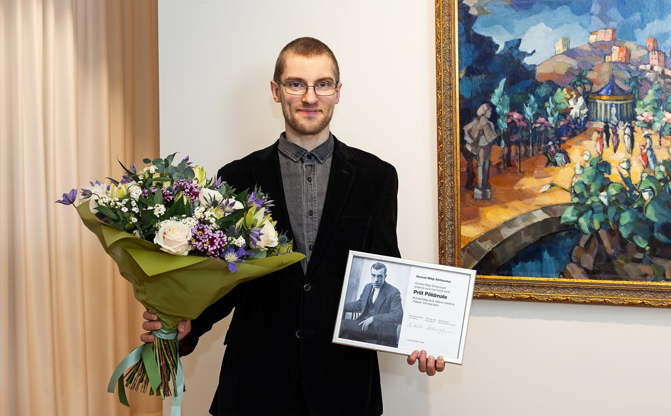 Pildil on Priit Põldma, kes hoiab ühes käes suurt lillekimpu ja teises Konrad Mäe pildiga diplomit. Tema selja taga paistab maal.