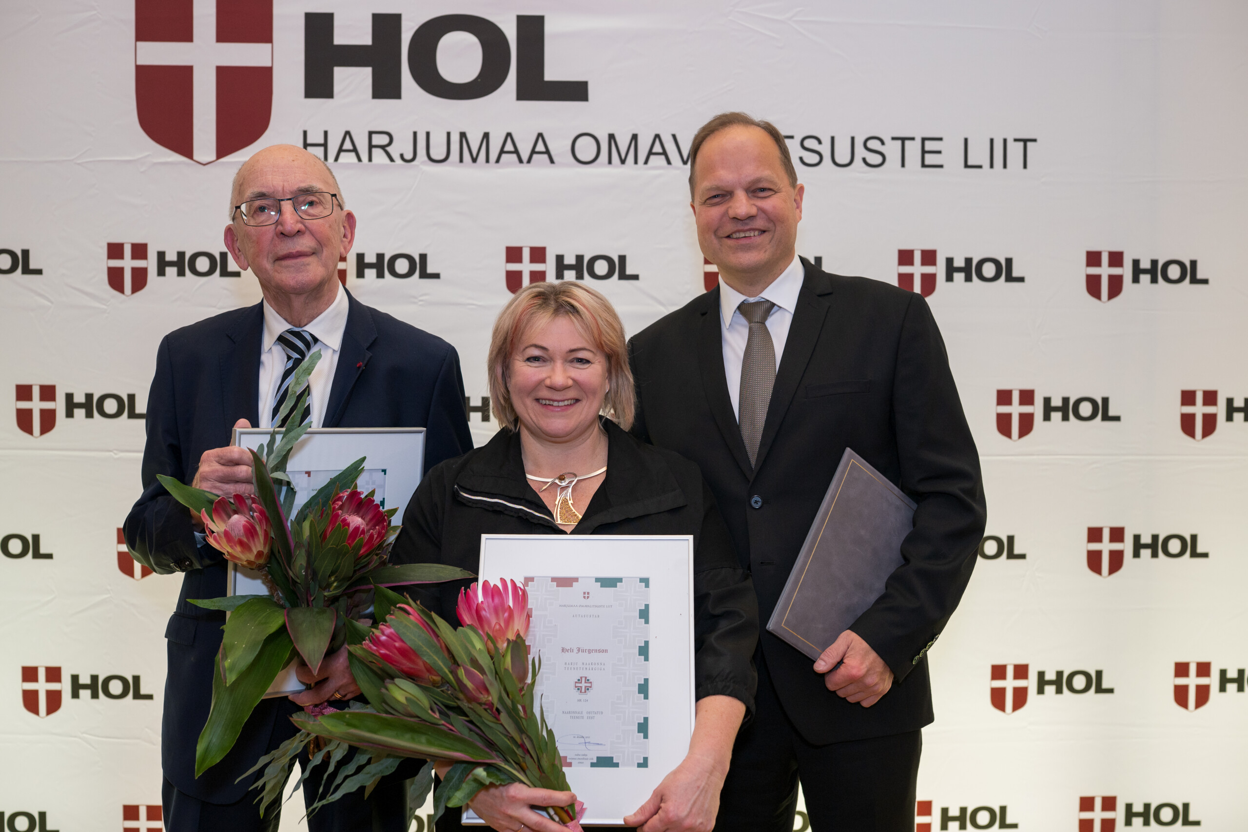 Pildil on Arne Mikk, Heli Jürgenson ja Ott Maaten, kes hoiavad käes diplome ja lilli. Nende taga paistab Harjumaa Omavalitsuste Liidu logodega sein.
