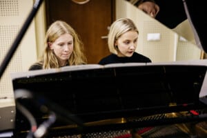 Pildil on Emili Rohumaa ja Hele Palumaa, kes istuvad helistuudios klaveri taga. Rohumaal on pikad heledad lokkis juuksed, Palumaal lühikesed heledad juuksed ja seljas must särk.