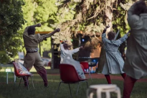 Pildil on neli etendajat, kes tantsivad pargis ja on pildistamise hetkel seljaga kaamera poole, käed üles tõstetud. Nende vahel paistavad punased plastiktoolid.