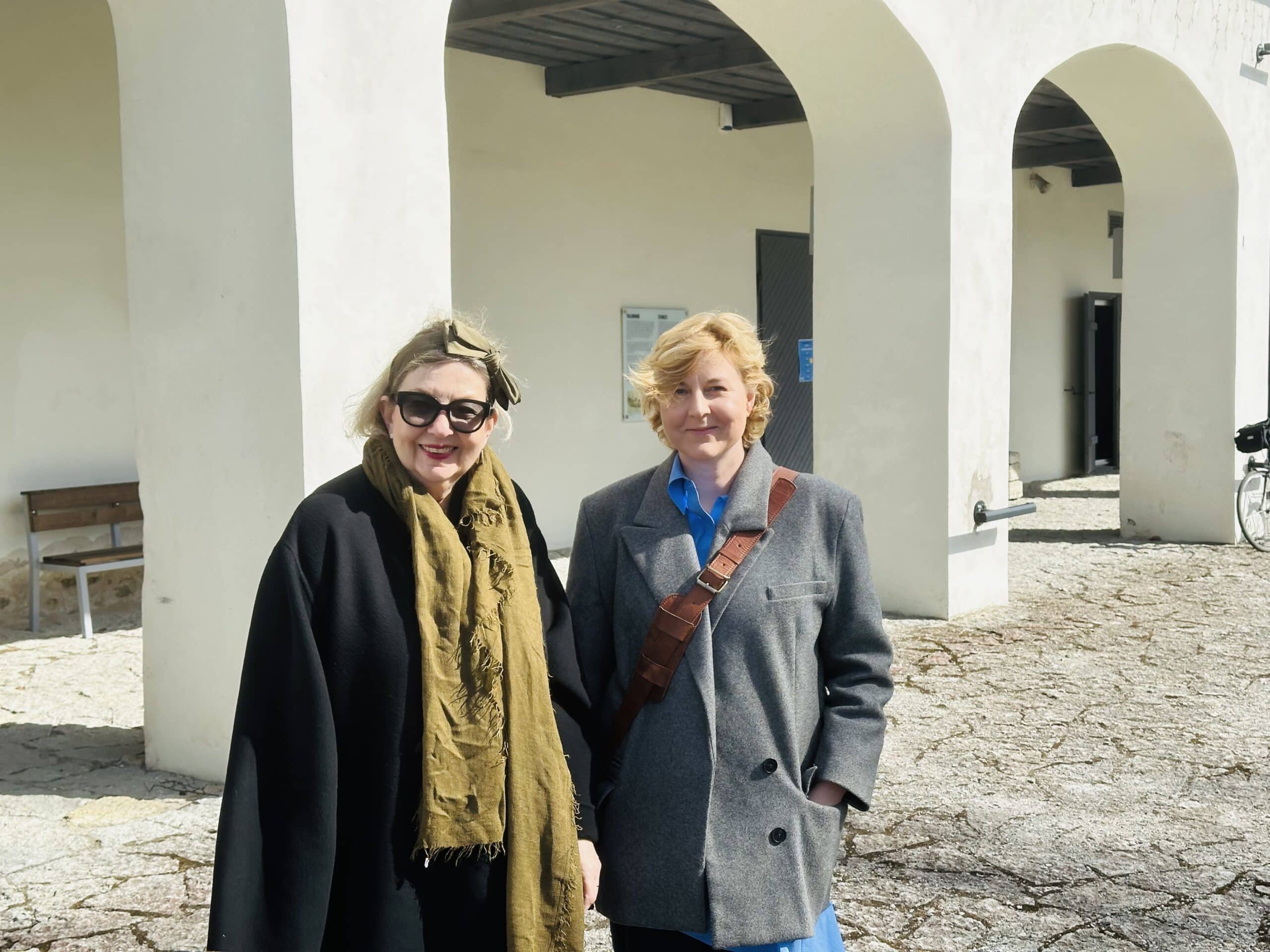 Pildil seisavad Lilja Blumenfeld ja Laura Pählapuu valgete sammastega tallihoone ees. Blumenfeldil on seljas must mantel ja rohekas sall, Pählapuul hall mantel.