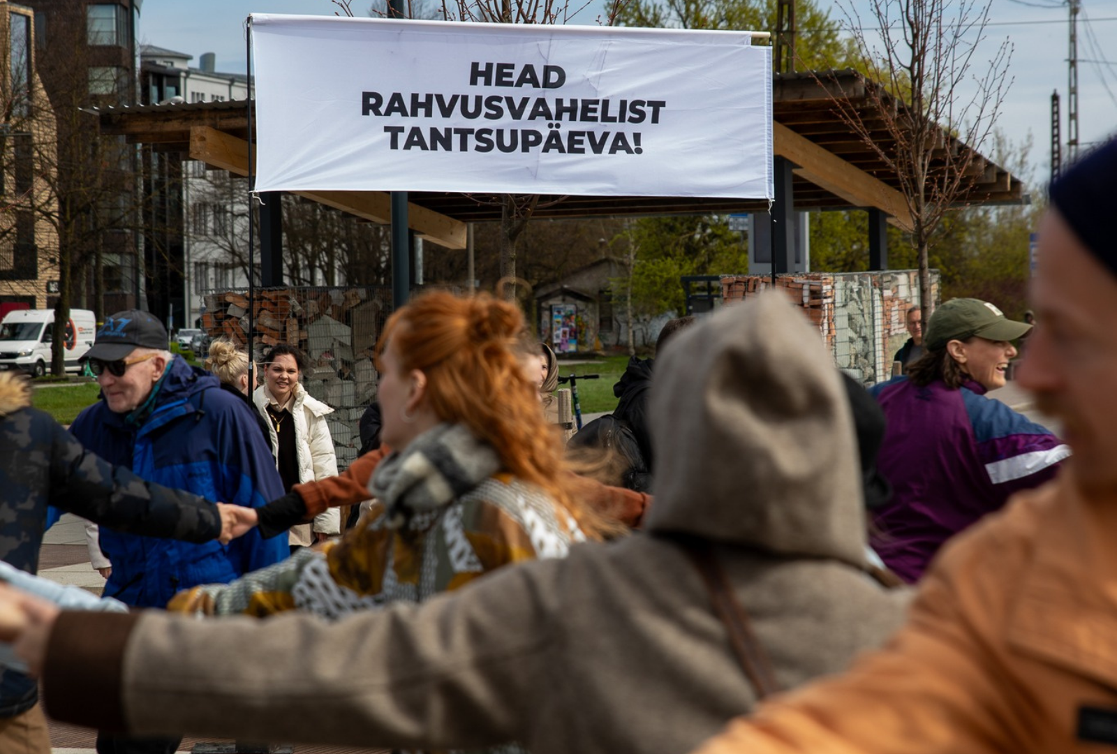 Pildil on näha avalikus ruumis liikuvaid inimesi, nende taga paistab suur valge silt musta kirjaga 