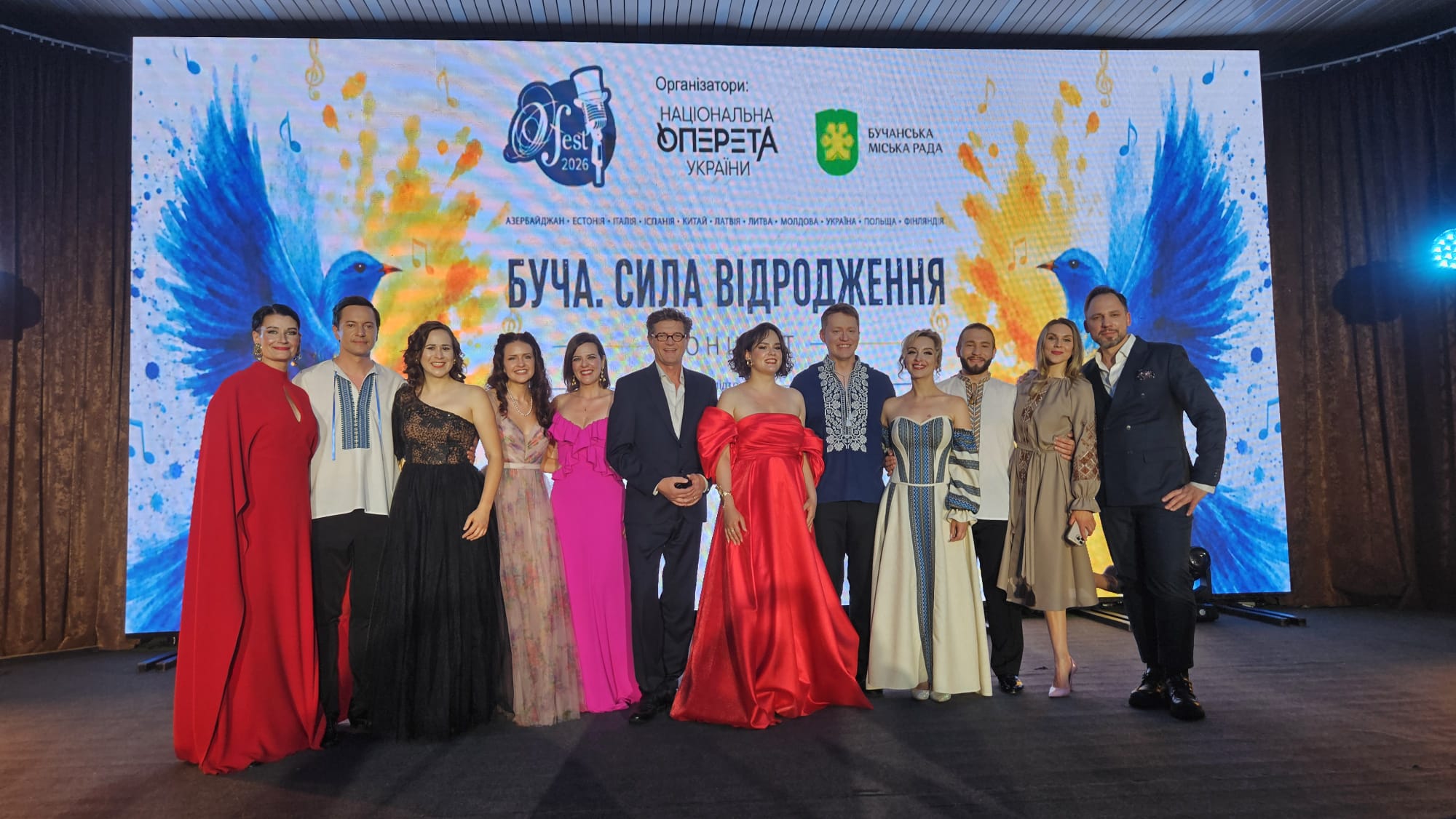 Pildil on 12 pidulikes riietes inimest, kes seisavad suure ekraani ees. Ekraanil on ukraina keeles kirjas "Butša. Taassünni jõud".