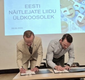 Pildil on Arvi Tavast ja Reimo Sagor, kes kummarduvad laua kohale, et kirjutada kokkuleppele allkirja. Nende taga paistab seinale projitseeritud slaid pealkirjaga 
