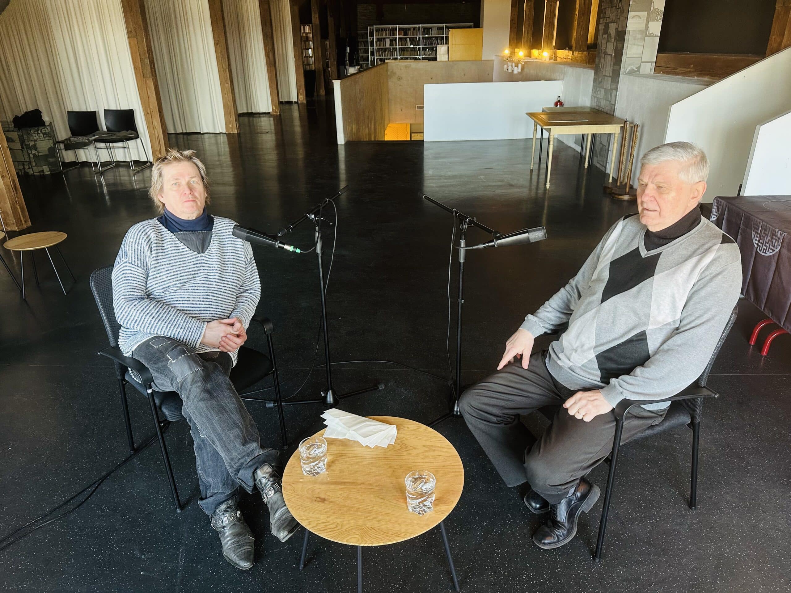 Pildil istuvad Mats Õun ja Priit Vaher toolidel, nende keskel väike puitlaud kahe veeklaasi ja salvrätikutega. Mõlema mehe ees on statiiviga mikrofon.