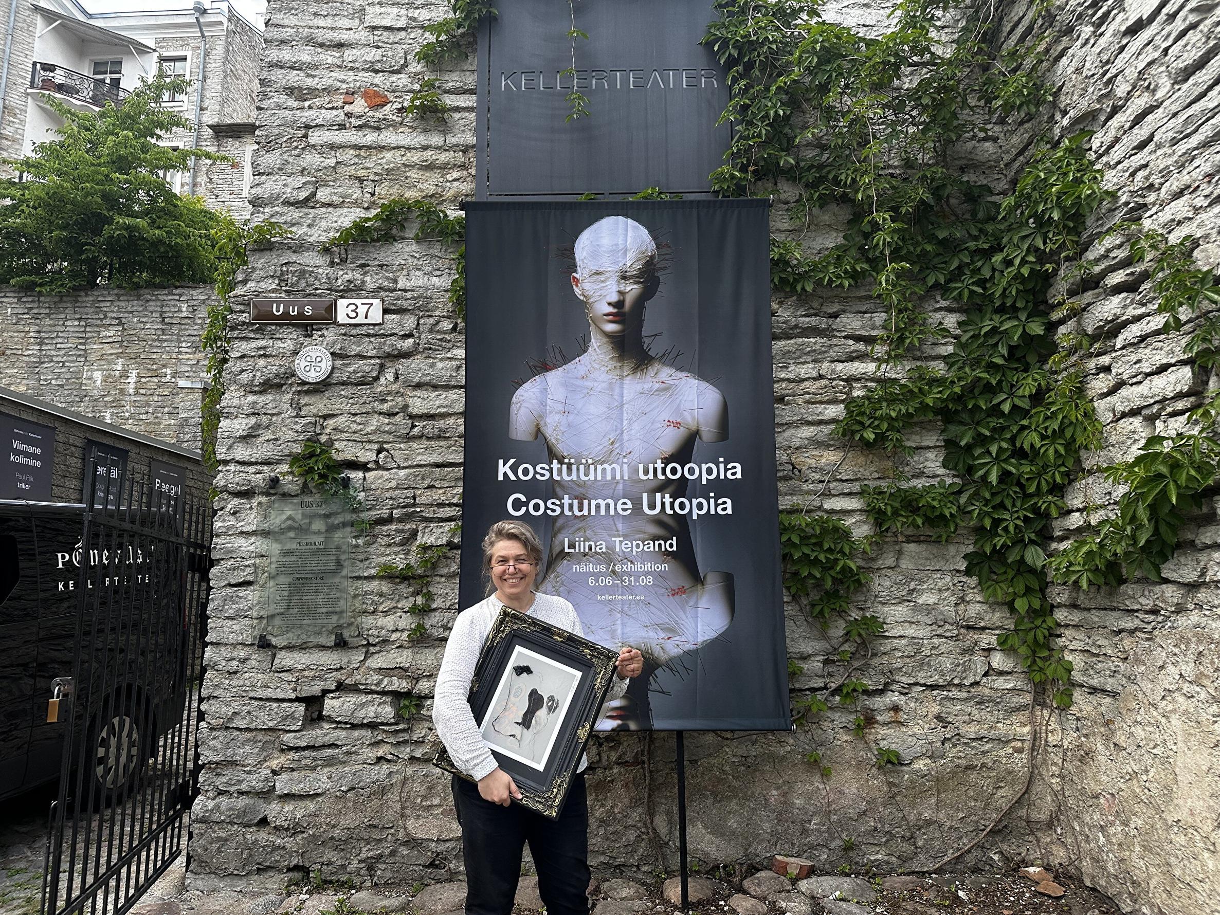 Pildil on Liina Tepand, kes seisab Püssirohuaida kiviseina ees. Tema selja taga paistab suur plakat kirjaga 