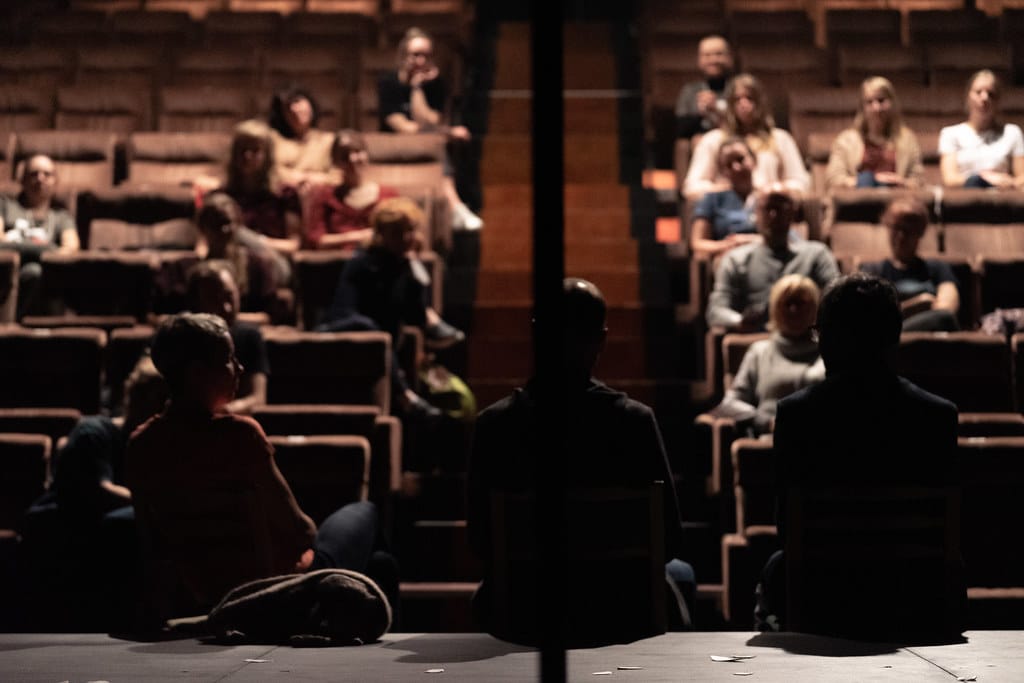 Pildil on Tartu Uue Teatri saal, kus istub paarkümmend inimest. Nende ees istub lava serval kolm vestlejat.