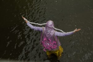 Esileht Pildil on näha etendajat, kes seisab jalgupidi vees ja seljaga kaamera poole. Ta hoiab käsi kahel pool laiali ja kannab seljas helelillat kostüümi.