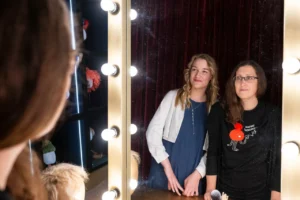 Pildil on Carolin Krajnak ja Annely Kaldoja, kes vaatavad kõrvuti seistes peeglisse. Peegel on ääristatud eredate lampidega.