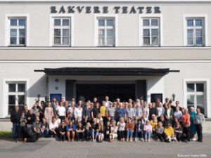 Pildil on mitukümmend inimest, kes seisavad ja istuvad Rakvere Teatri suure maja peaukse ees. Nende taga paistab helehall sein, uks ja silt 