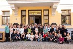 Pildil seisab ja kükitab mitukümmend inimest Eesti Noorsooteatri Laia tänava poolse sissepääsu ees. Nende taga on näha helekollast seina, ust ja silti 