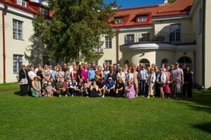 Pildil on mitukümmend inimest, kes seisavad ja kükitavad muru peal, taustal helekollaste seinte ja punase katusega hoone.