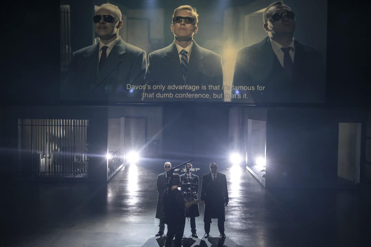 Pildil on stseen lavastusest "Rahamaa", kus kolm näitlejat seisavad eeslaval ja nende ees operaator. Nende selja taga on projitseeritud operaatori filmitav live-video.